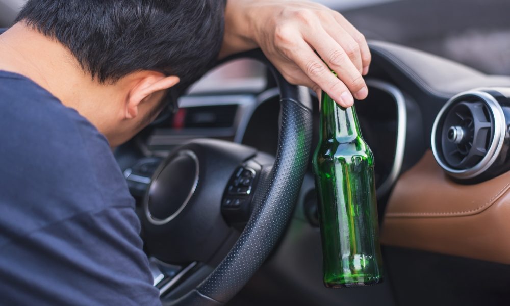 A young man leaning over the steering wheel of an automobile, lazily holding a green bottle of beer in his hand.