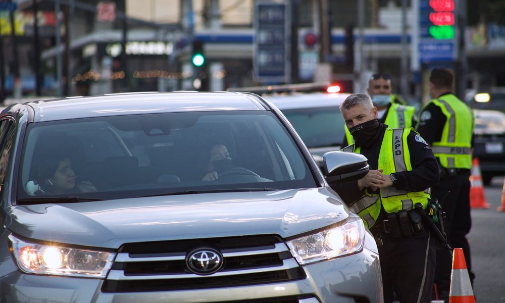 A masked police officer at a DUI checkpoint in California conversing with a driver.