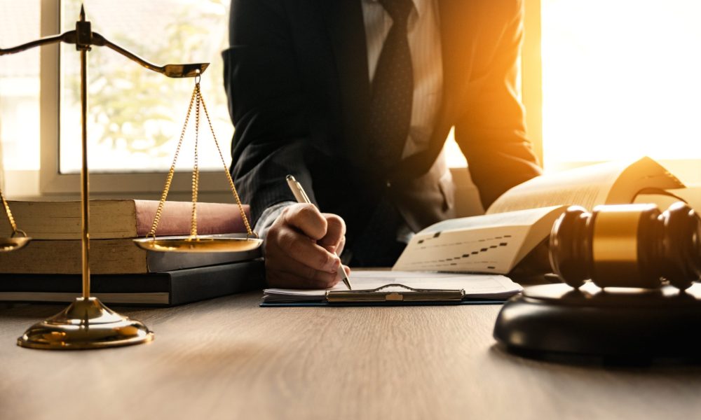 A man writing in a notebook at a desk, a balance beside his right hand with a gavel resting on the desk.