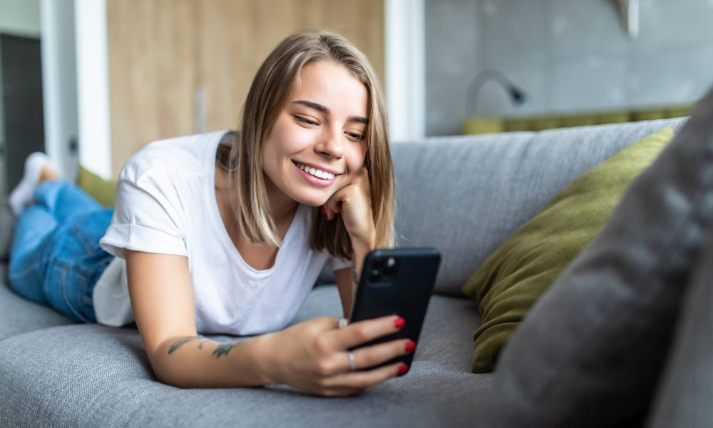 A woman smiling and laying on a coach while using her smart phone.